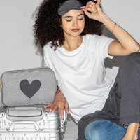 Woman wearing a grey cotton eye mask, preparing for a trip with a silver suitcase and a heart-shaped travel blanket pouch.