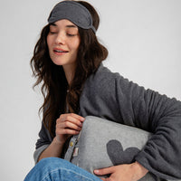 Woman wearing a gray sleep mask and holding a matching heart-shaped travel blanket pouch.