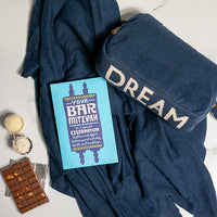 Navy blue travel blanket with the word 'dream' on it, Bar Mitzvah card, chocolate, and cupcakes on a light background.