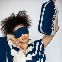 Woman wearing a blue cotton sleep mask and holding a navy blue and white bag.