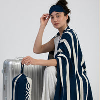 Woman wrapped in a striped navy and white blanket holding a silver suitcase with a blue eye mask on her head against a white background