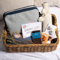 Jute gift basket with a gray travel blanket pouch, cards, chocolate, knit wine pouch and a plant on a white surface.
