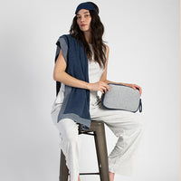 Woman wearing a blue eye mask and holding a gray travel pouch with a blue cotton blanket draped over her shoulders on a white background
