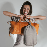 Woman smiling and holding a brown leather bag and a gray fabric pouch against a plain background.