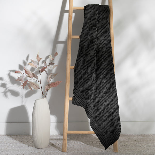 Black charcoal patterned chunky knit blanket draped over a wooden ladder against a white wall with a vase of leaves beside it.