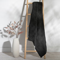 Black charcoal patterned chunky knit blanket draped over a wooden ladder against a white wall with a vase of leaves beside it.