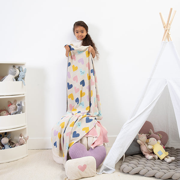 Child holding a colorful heart-patterned blanket in a room with toys and a teepee.