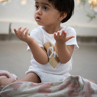 Child wearing a white shirt with a heart design, sitting on a cotton blanket with pink bunny design.