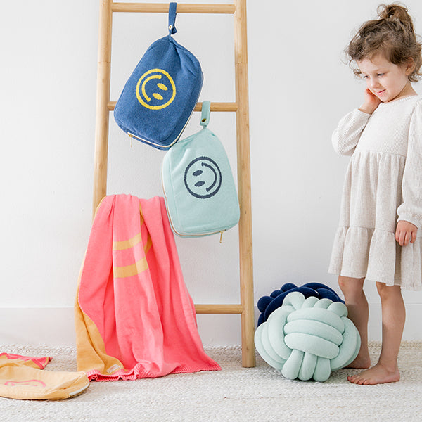 Child standing next to a wooden ladder with colorful bags and a two knotted cushions on a light-colored floor.