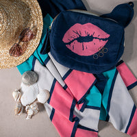 Navy blue pouch with pink lip design, straw hat, sunglasses, and travel blanket on a neutral surface