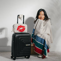 Woman wrapped in a beige striped blanket next to a suitcase with a small bag featuring red lips on a plain background