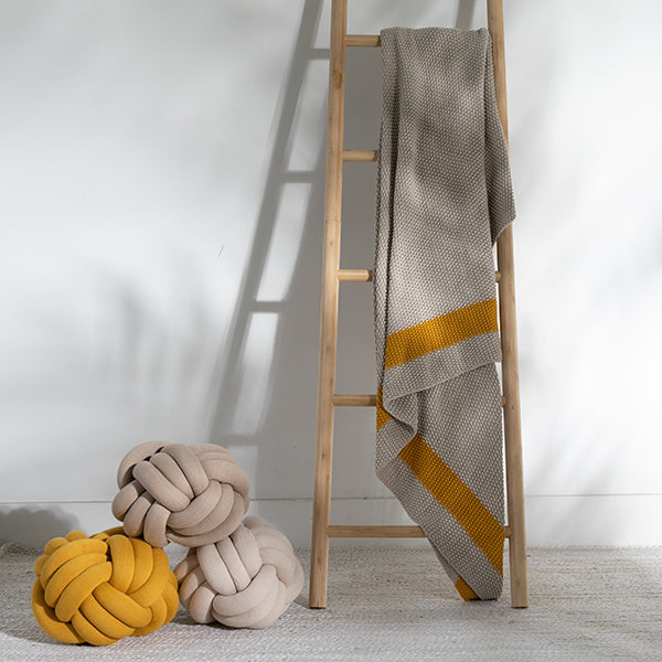 Yellow and white knitted cotton ball cushions and blanket on a wooden ladder against a white wall.