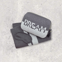 Gray organic cotton travel pouch with 'DREAM' text, dark gray eye mask, and gray blanket with light grey stripes on a light gray textured background.
