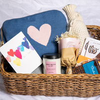 A variety of items including a blue and pink reversible blanket set, a zip pouch, a wristlet loop, a sleep mask, a candle, a card with a heart design, displayed in a wicker basket.
