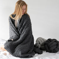Woman wrapped in a dark gray cable knit blanket with additional pillows on a white background.