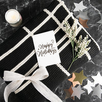 Gift wrapped black and white moss-knit throw blanket with a 'Happy Holidays' card on a dark surface with decorative stars.