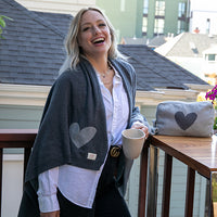 Woman smiling on a balcony wrapped with a grey travel blanket, holding a coffee mug next to a table with a grey heart travel pouch.