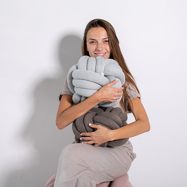Woman holding two large knitted pillows in blue and dark grey against a plain background.