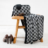A black and gray geometric patterned travel blanket and sleep mask set, displayed on a wooden stool next to high heels.