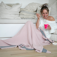 Woman sitting next to a white couch with a pink moss stitch knit cotton blanket, eating ice-cream from a pink cup.