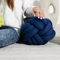 Navy blue knot pillow on a light gray surface with a person sitting nearby.