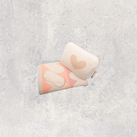 A cotton travel blanket case with a heart design with a matching beige eye mask and blanket on a light gray textured background.