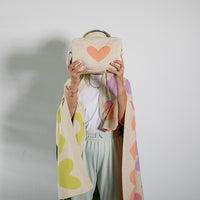 Person holding a beige bag with a heart design and wearing a blanket against a plain background.