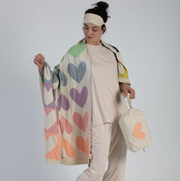 Woman wearing an eye mask and holding a colorful heart-patterned blanket and blanket bag against a plain background.
