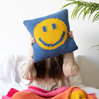 Person holding a blue knitted cotton cushion with a yellow smiley face design in front of their face while sitting on a bed.