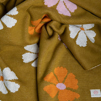 Close up of a floral patterned throw blanket with white, orange, and pink flowers on a bush green background.
