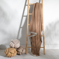 Linen brown woven cotton blanket draped over a wooden ladder with three knot-shaped cushions on the floor.