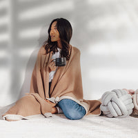 Woman wrapped in a linen textured blanket holding a mug, sitting on a white surface with a white knot pillow.