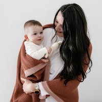 Woman holding a baby wrapped in a rust cotton knit blanket against a white background.