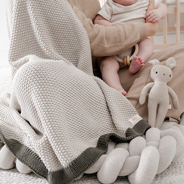 Person holding a baby wrapped in a knitted lekha sand blanket with a bunny soft toy on a bed.