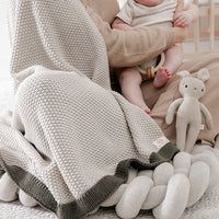 Person holding a baby wrapped in a knitted lekha sand blanket with a bunny soft toy on a bed.
