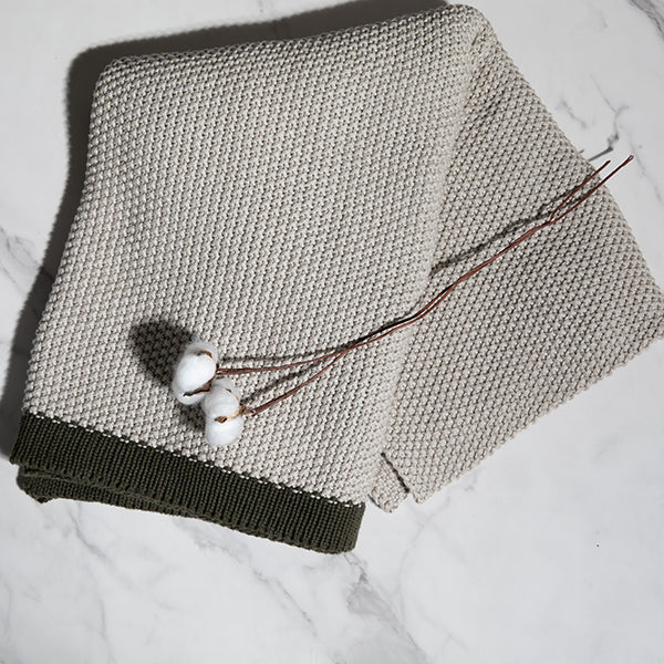 Folded cotton knit throw blanket with a dark border with a decorative plant on a marble surface.