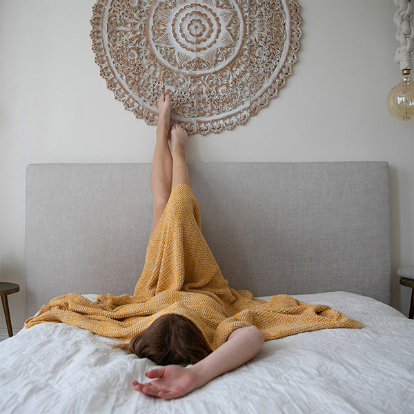 Person lying on a bed with a yellow blanket, feet raised, against a decorative wall mandala.