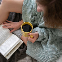 Person reading a book and holding a yellow mug with a blue throw blanket wrapped around them.