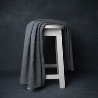 Chunky knitted light grey organic cotton throw blanket draped over a white stool against a dark background.
