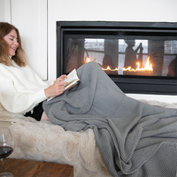 Woman enjoying a book on a couch next to a warm fireplace with a gray chunky knitted throw.