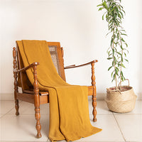 Yellow knitted cotton throw blanket draped over a wooden and jute chair next to a tall plant in the background.