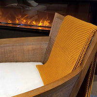 Mustard yellow chunky knitted cotton throw blanket draped over a jute chair in front of a fireplace.