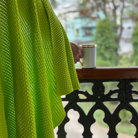 Bright neon yellow knitted blanket draped around a person standing on a balcony overlooking trees while holding a white mug.