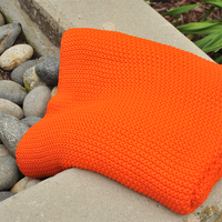 Folded bright orange knitted throw on a stone surface with a natural background next to plants and pebbles.