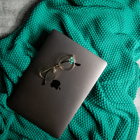 Black Apple macbook with a pair of glasses placed on a sea green textured cotton blanket.