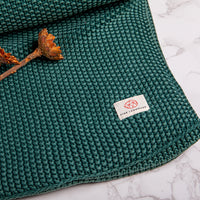 Emerald green textured knit cotton throw blanket with a Pink Lemonade brand label and dried plants on a marble surface.