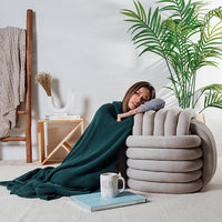 Woman wrapped in a emerald green knit throw laying next to a large grey knot cushion with a plant and books in the background.
