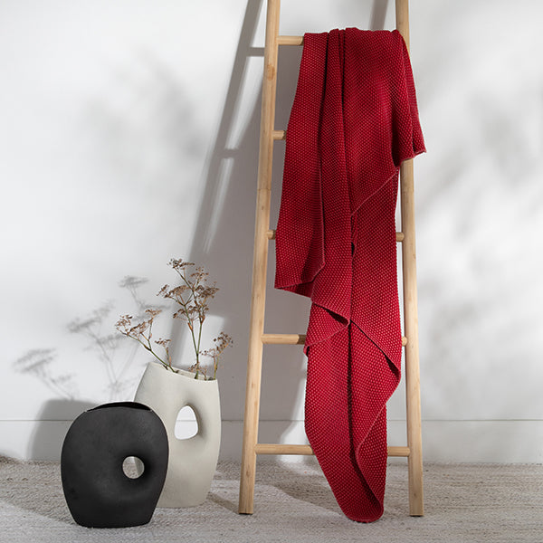 Red knit cotton blanket draped over a wooden ladder against a white wall with a vase and decorative item on the floor.