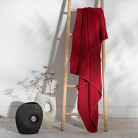 Red knit cotton blanket draped over a wooden ladder against a white wall with a vase and decorative item on the floor.