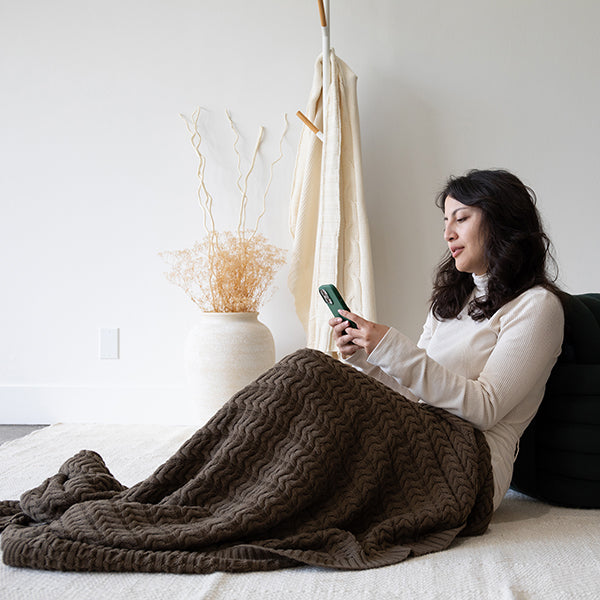 Woman sitting on a bed using a phone with a dark green braided knit cotton throw draped over her, in a room with white walls and decor.